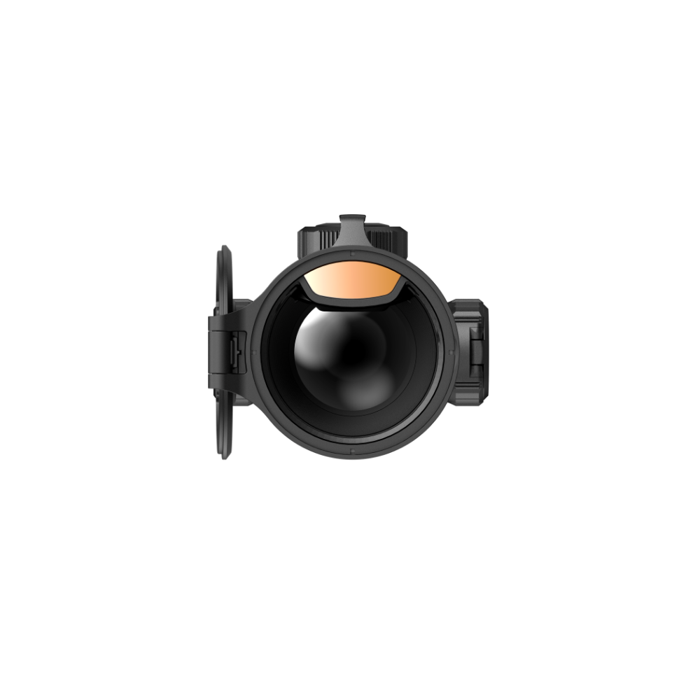 A black tactical rifle scope with a lens cover with a hinged side flap, shown from the front. The lens inside has a warm orange-tinted insert visible above the main black lens.